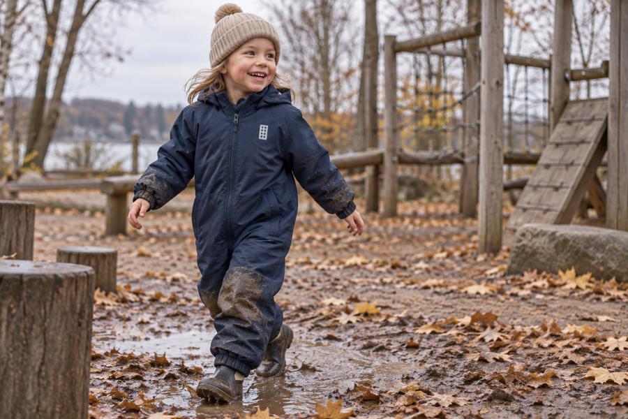 Happy five-year-old child in a muddy dark navy LWJORI 721 snowsuit at a playground in late autumn.
