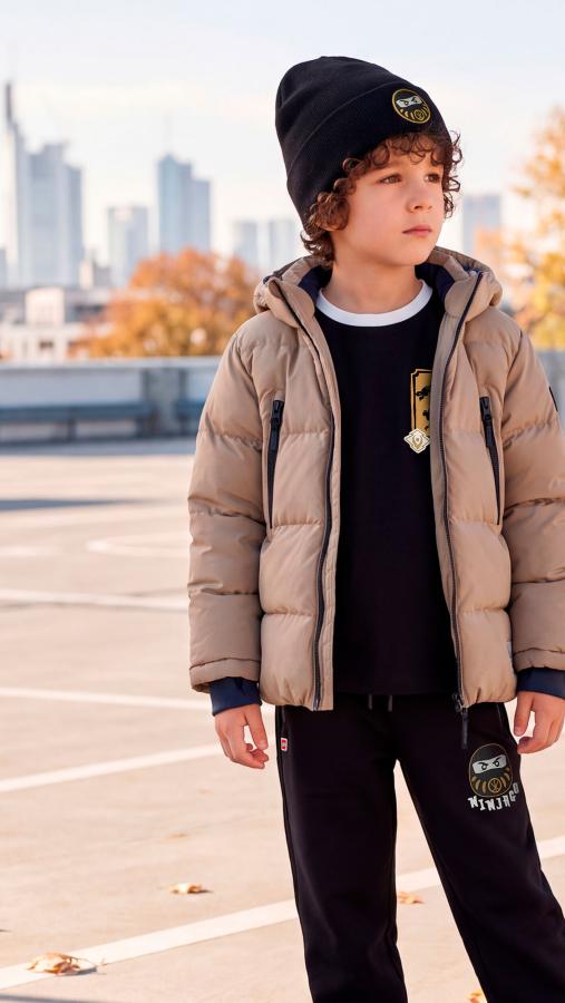 Child in a beige winter jacket and black LEGO® NINJAGO® hat on an urban rooftop
